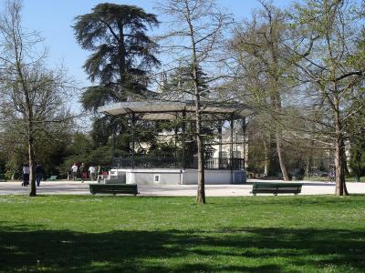 A metal and concrete bandstand (kiosk) situated in a grassy public park with trees, benches, and people walking in the background under a clear blue sky.