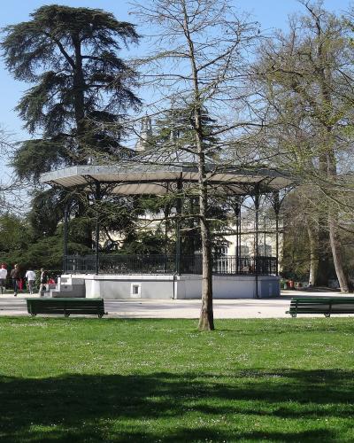 A metal and concrete bandstand (kiosk) situated in a grassy public park with trees, benches, and people walking in the background under a clear blue sky.
