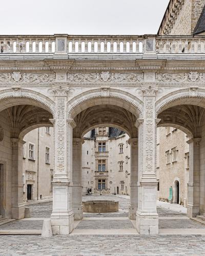 The image shows the ornate stone arcade of the Cour d'Honneur (Courtyard of Honor) at the Château de Pau, featuring three large arches with intricate Renaissance carvings and royal monograms, leading into an inner courtyard with a fountain.