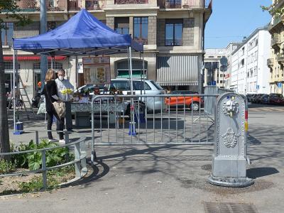 The image shows an outdoor street scene with a blue canopy tent set up on a paved area, where two people are interacting near a table; in the foreground, there is a distinctive grey stone public drinking fountain (borne-fontaine) with sculpted det