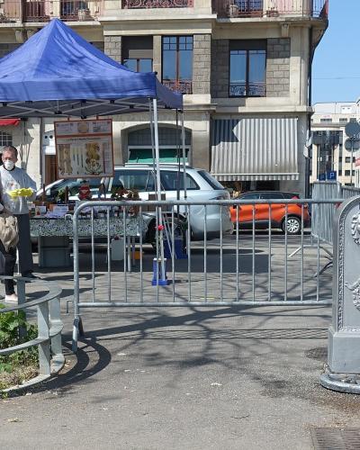 The image shows an outdoor street scene with a blue canopy tent set up on a paved area, where two people are interacting near a table; in the foreground, there is a distinctive grey stone public drinking fountain (borne-fontaine) with sculpted det