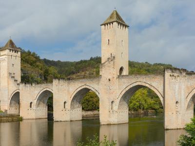 A stone medieval fortified bridge with three prominent towers and multiple arches spanning a river, set against a backdrop of green hills under a cloudy sky.