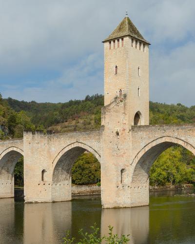 A stone medieval fortified bridge with three prominent towers and multiple arches spanning a river, set against a backdrop of green hills under a cloudy sky.