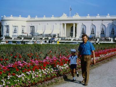 A large, white, neoclassical building with a curved facade, columns, and statues on the roofline, viewed from across a gravel path lined with colorful flowerbeds; a man and a young boy in sailor-style clothing walk in the foreground, and vintage c