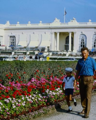 A large, white, neoclassical building with a curved facade, columns, and statues on the roofline, viewed from across a gravel path lined with colorful flowerbeds; a man and a young boy in sailor-style clothing walk in the foreground, and vintage c