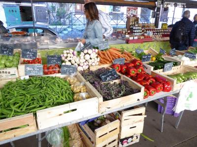 A street market stall displaying fresh produce like green beans, carrots, peppers, and onions in wooden crates with handwritten French price signs.