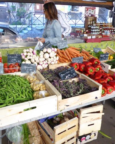 A street market stall displaying fresh produce like green beans, carrots, peppers, and onions in wooden crates with handwritten French price signs.
