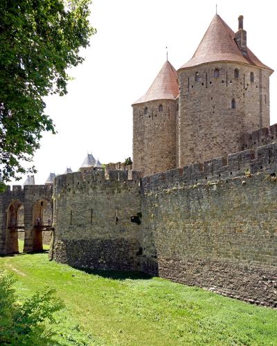 The image shows the stone ramparts and conical-roofed towers of the fortified city of Carcassonne, specifically the section near the Narbonne Gate (Porte Narbonnaise), with a grassy moat in the foreground.