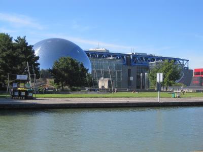 The image shows the exterior of the Cité des Sciences et de l'Industrie in Paris, featuring its iconic silver geodesic dome (La Géode) and the blue steel structure of the Grande Halle, viewed across a canal in Parc de la Villette.
