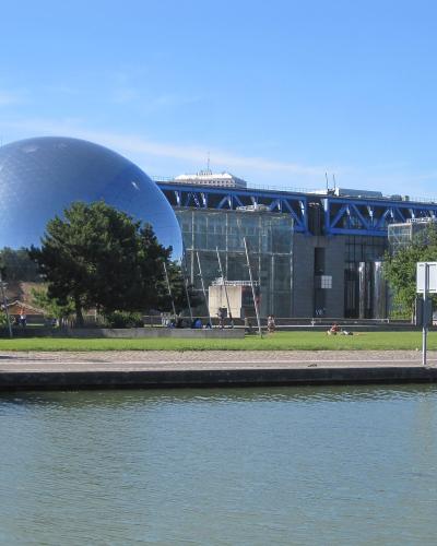 The image shows the exterior of the Cité des Sciences et de l'Industrie in Paris, featuring its iconic silver geodesic dome (La Géode) and the blue steel structure of the Grande Halle, viewed across a canal in Parc de la Villette.