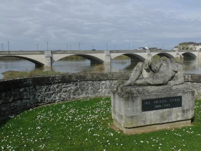 A stone war memorial sculpture of a reclining figure on a grassy bank with white flowers, overlooking a multi-arched concrete bridge spanning a wide river under an overcast sky.