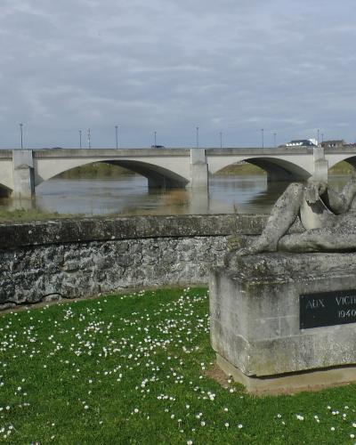 A stone war memorial sculpture of a reclining figure on a grassy bank with white flowers, overlooking a multi-arched concrete bridge spanning a wide river under an overcast sky.
