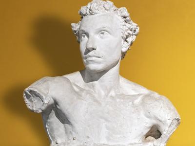 A white plaster or terracotta bust of a man with curly hair and a mustache, missing its right arm, displayed against a solid yellow wall on a marble-like pedestal.