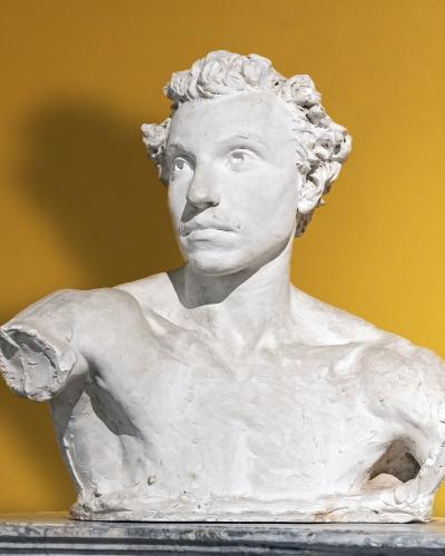 A white plaster or terracotta bust of a man with curly hair and a mustache, missing its right arm, displayed against a solid yellow wall on a marble-like pedestal.