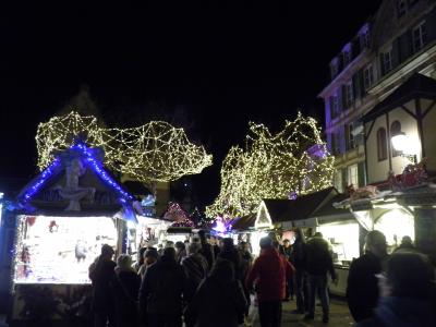 A night scene of a crowded outdoor Christmas market with wooden stalls, festive string lights, and people in winter clothing.