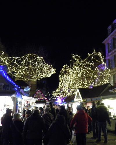 A night scene of a crowded outdoor Christmas market with wooden stalls, festive string lights, and people in winter clothing.