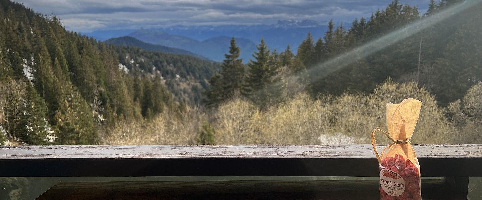 1. A view from a wooden balcony or railing overlooking a forested mountain valley with snow-dusted slopes and distant peaks under a cloudy sky, with a small bag of dried fruit labeled "Fruits de Genièvre" in the foreground.