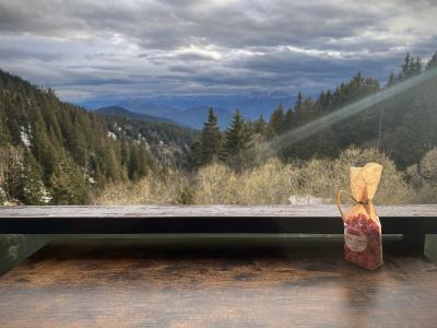 1. A view from a wooden balcony or railing overlooking a forested mountain valley with snow-dusted slopes and distant peaks under a cloudy sky, with a small bag of dried fruit labeled "Fruits de Genièvre" in the foreground.