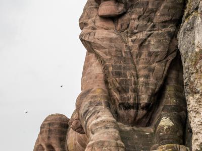 A low-angle photograph of the massive Lion of Belfort statue carved into the sandstone cliff face, showing its head, front paws, and part of its body against an overcast sky.