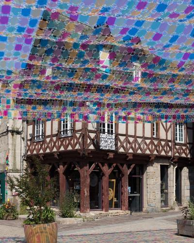 The image shows a pedestrian square in a French town featuring a prominent half-timbered building and a large, colorful canopy of plastic bottles suspended overhead.