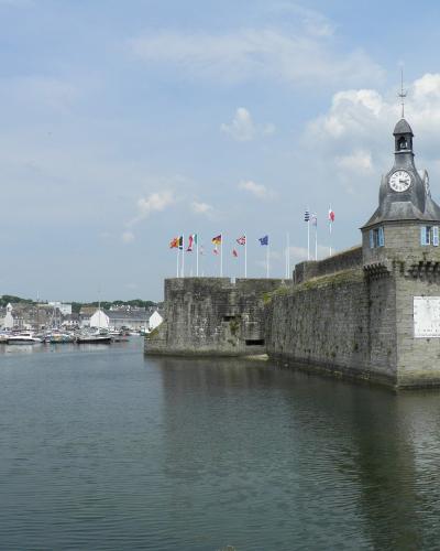 A stone fortification with a clock tower and multiple flags stands at the entrance of a harbor filled with boats, with a town visible in the background.