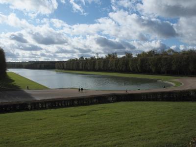 Wide-angle view of the Grand Canal in Versailles Gardens with visitors strolling along the banks under a cloudy sky.