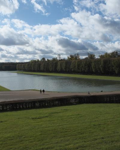 Wide-angle view of the Grand Canal in Versailles Gardens with visitors strolling along the banks under a cloudy sky.