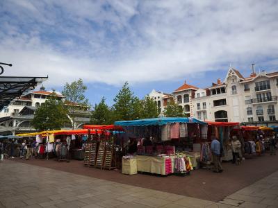 An outdoor market with colorful stalls selling clothing and goods, set on a paved plaza with distinctive white buildings featuring red-tiled roofs and ornate balconies in the background under a partly cloudy sky.