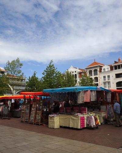 An outdoor market with colorful stalls selling clothing and goods, set on a paved plaza with distinctive white buildings featuring red-tiled roofs and ornate balconies in the background under a partly cloudy sky.