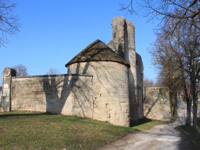 The image shows the stone ruins of a medieval fortification, specifically a round tower with a conical roof and an adjacent rectangular tower, situated behind a stone wall with a metal gate on the left, under a clear blue sky.