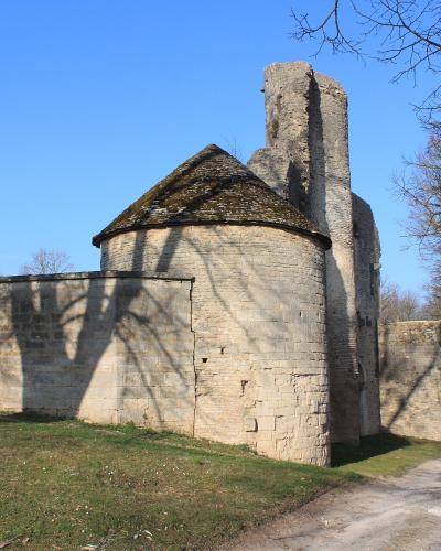 The image shows the stone ruins of a medieval fortification, specifically a round tower with a conical roof and an adjacent rectangular tower, situated behind a stone wall with a metal gate on the left, under a clear blue sky.