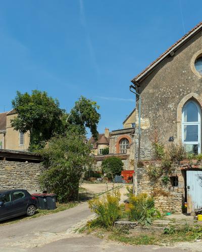 A sunlit street scene in a French village featuring a weathered stone building with Gothic-arched windows and a round oculus, alongside a parked car and stone walls under a clear blue sky.