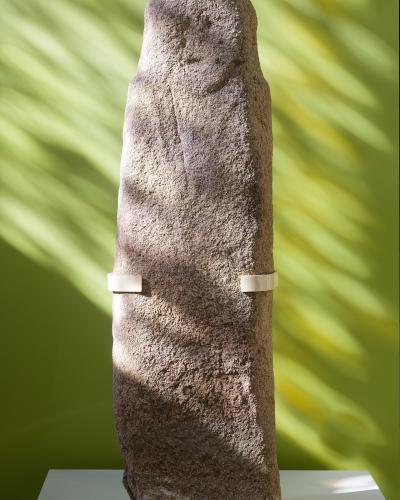A tall, weathered stone statue-menhir displayed on a white pedestal against a green wall with dappled light shadows; two white conservation brackets are attached to its midsection.