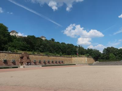 A large outdoor memorial featuring a long stone wall with bronze reliefs and a central cross, situated on a hillside with a flagpole and a large paved plaza in front.