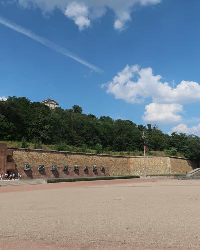 A large outdoor memorial featuring a long stone wall with bronze reliefs and a central cross, situated on a hillside with a flagpole and a large paved plaza in front.