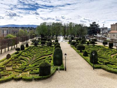 A formal French garden with intricate boxwood parterres, gravel paths, sculpted topiary, and a central fountain, viewed from an elevated position with urban buildings and distant hills in the background; signage or text not visible in image.