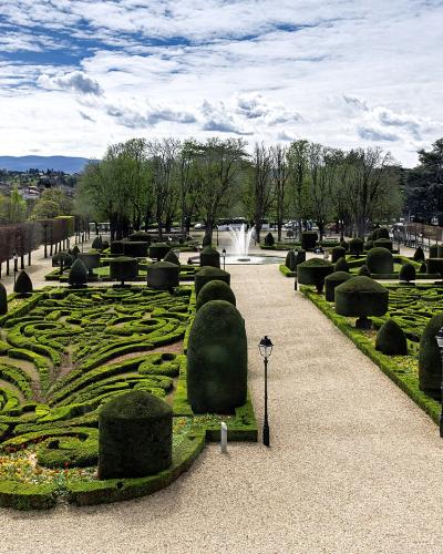 A formal French garden with intricate boxwood parterres, gravel paths, sculpted topiary, and a central fountain, viewed from an elevated position with urban buildings and distant hills in the background; signage or text not visible in image.