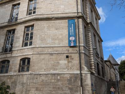 A low-angle exterior shot of a historic stone building with a blue vertical banner reading "MUSÉE DE LA CÉRAMIQUE" hanging on its facade, alongside a parked car and a person on stairs.
