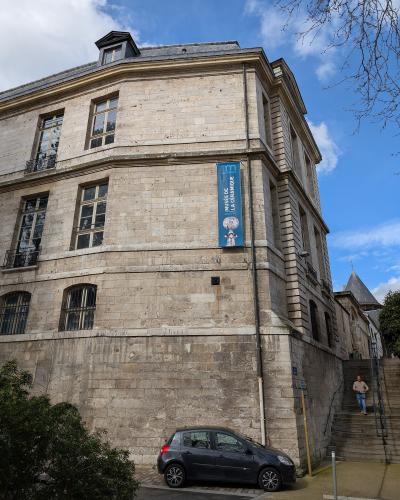 A low-angle exterior shot of a historic stone building with a blue vertical banner reading "MUSÉE DE LA CÉRAMIQUE" hanging on its facade, alongside a parked car and a person on stairs.