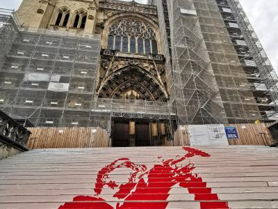 A low-angle view of a Gothic cathedral facade covered in scaffolding and protective netting, with a wide set of stairs in the foreground featuring a large red stencil artwork of a figure.