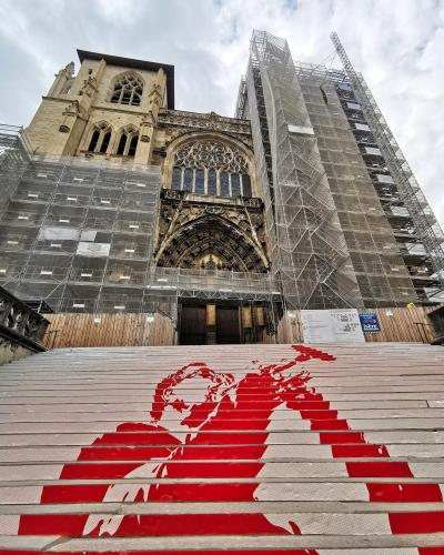 A low-angle view of a Gothic cathedral facade covered in scaffolding and protective netting, with a wide set of stairs in the foreground featuring a large red stencil artwork of a figure.