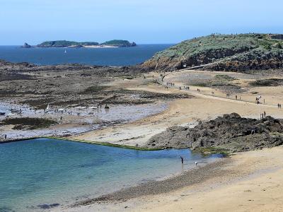 A coastal landscape showing a sandy beach with a rectangular tidal pool in the foreground, a rocky causeway leading to a grassy island with a stone fortification on the left, and distant islands under a clear blue sky; people are walking along the