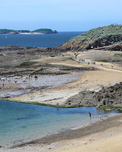 A coastal landscape showing a sandy beach with a rectangular tidal pool in the foreground, a rocky causeway leading to a grassy island with a stone fortification on the left, and distant islands under a clear blue sky; people are walking along the