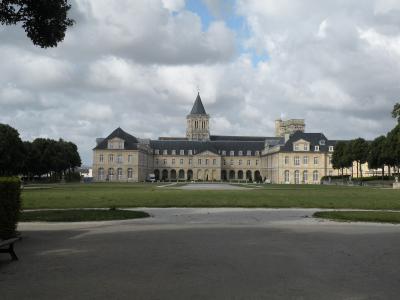 A wide-angle photograph of a large, historic stone monastery complex with a central bell tower and symmetrical wings, set behind a grassy lawn under a cloudy sky.