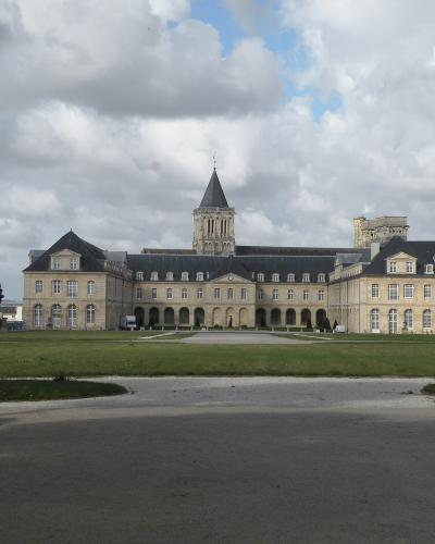 A wide-angle photograph of a large, historic stone monastery complex with a central bell tower and symmetrical wings, set behind a grassy lawn under a cloudy sky.