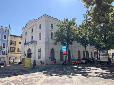 A white neoclassical building with arched windows and a pediment, identified by signage as "THÉÂTRE DE VALÈNE" and "VILLE DE VALÈNE", situated on a paved public square with trees and parked cars.