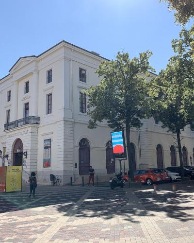 A white neoclassical building with arched windows and a pediment, identified by signage as "THÉÂTRE DE VALÈNE" and "VILLE DE VALÈNE", situated on a paved public square with trees and parked cars.