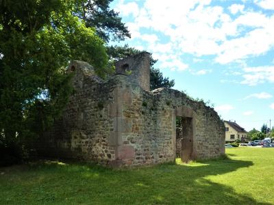 The image shows the stone ruins of a historic building, likely a former church or chapel, situated on a grassy lawn with large trees and a blue sky in the background.