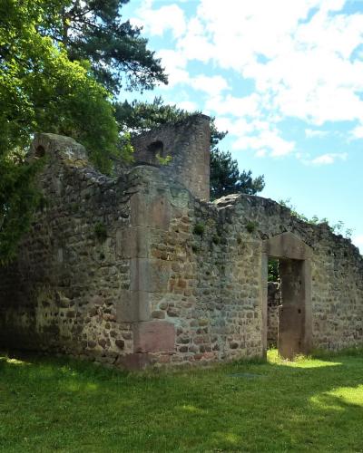 The image shows the stone ruins of a historic building, likely a former church or chapel, situated on a grassy lawn with large trees and a blue sky in the background.