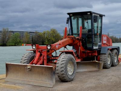 A red O&K motor grader parked on a dirt construction site next to a river, with a large modern building under construction visible in the background across the water.